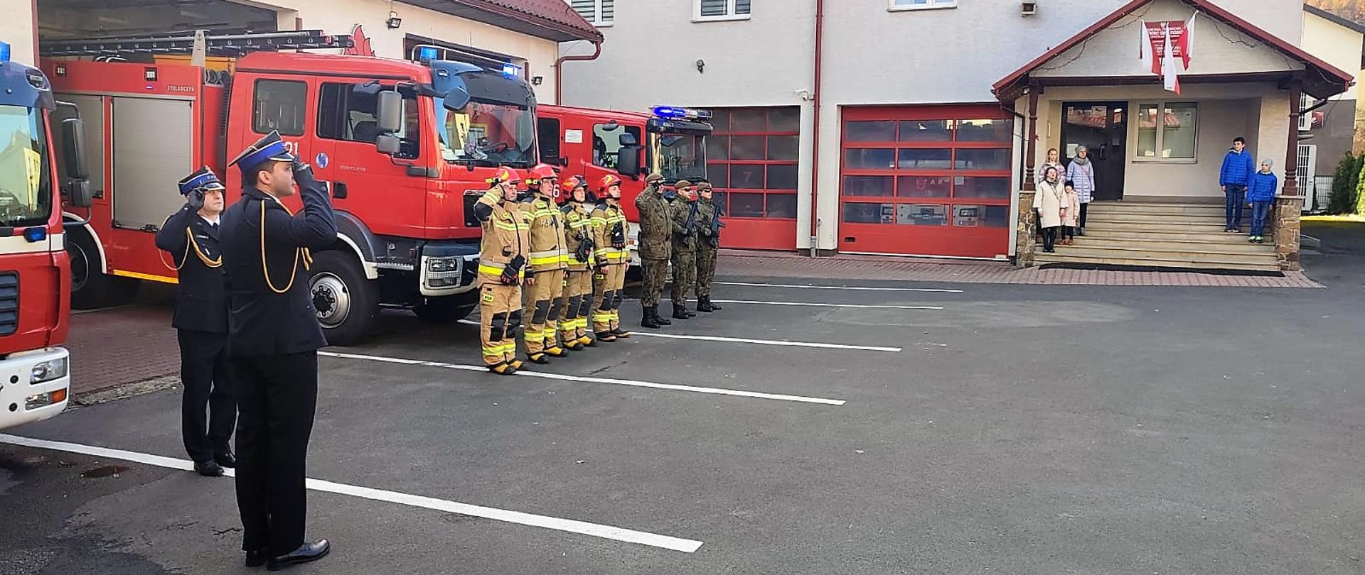 Zdjęcie wykonane zostało na placu Komendy Powiatowej PSP w Ustrzykach Dolnych. Widać na nim wozy bojowe które wystawione są z garaży i przed którymi stoją wojskowi, zmiana służbowa w ubraniach specjalnych oraz dwóch funkcjonariuszy PSP w mundurach galowych. Dowódcy oraz funkcjonariusze w mundurach galowych oddają honor. Na schodach głównego wejścia do komendy stoją osoby cywilne. 