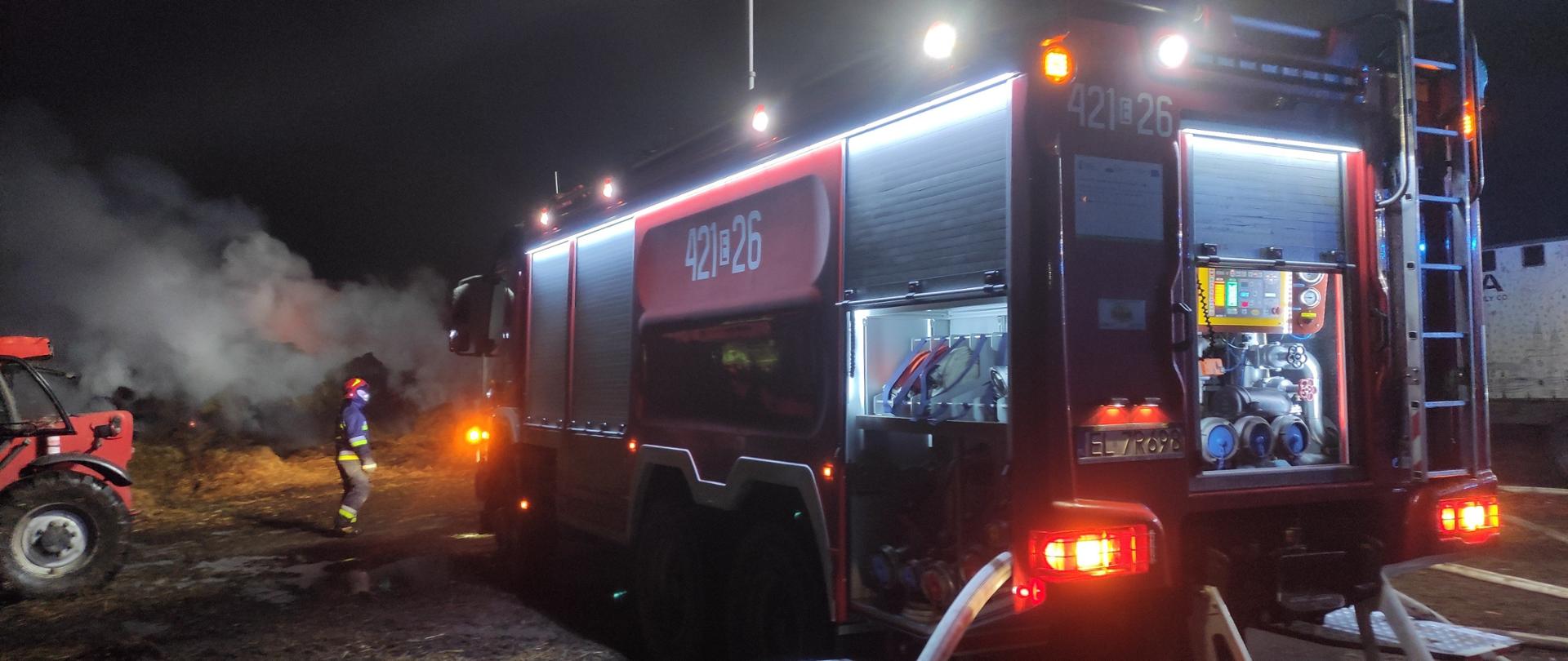 Wóz strażacki w czasie akcji gaśniczej oświetlony dookoła. W tle dym z palących się belek słomy. Na ziemi rozwinięta linia gaśnicza z węży strażackich.