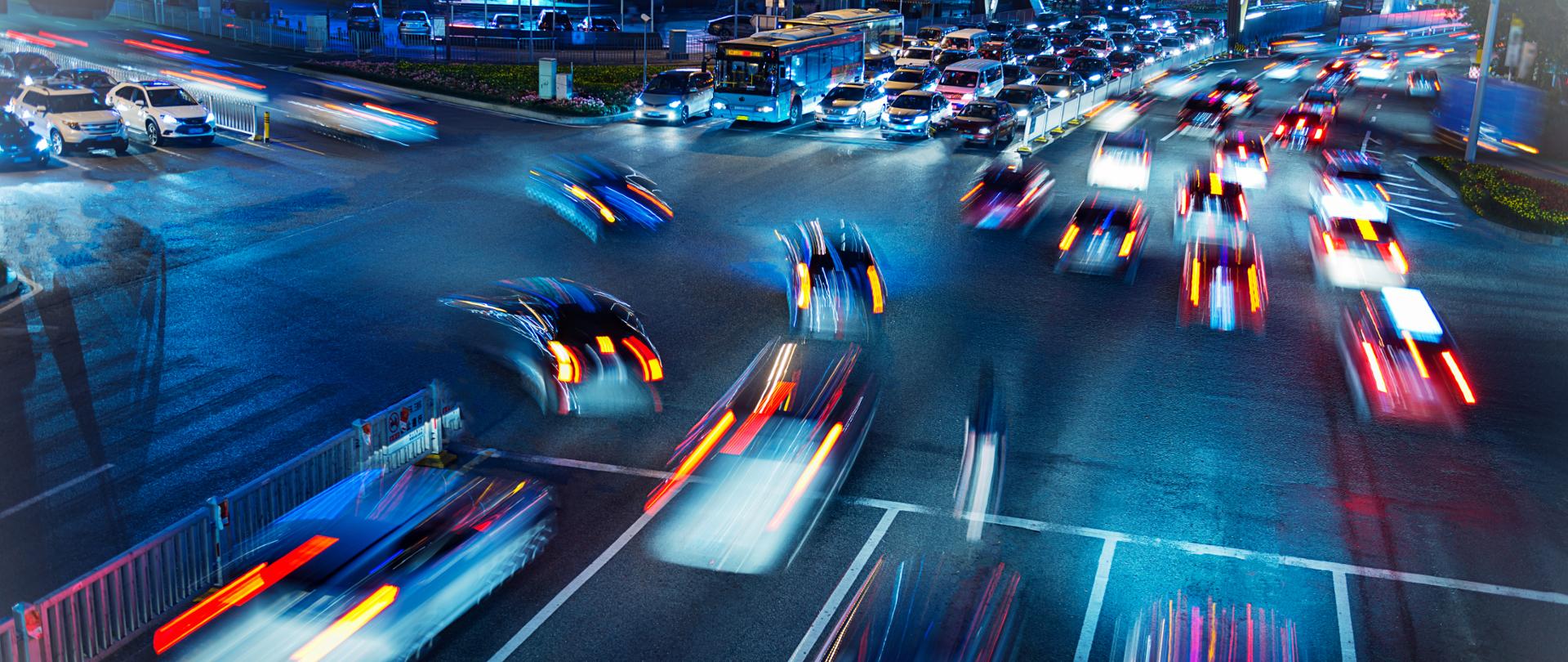Zdjęcie skrzyżowania drogowego (po zmroku) - widoczne kolorowe światła samochodów jadących w różne strony.
