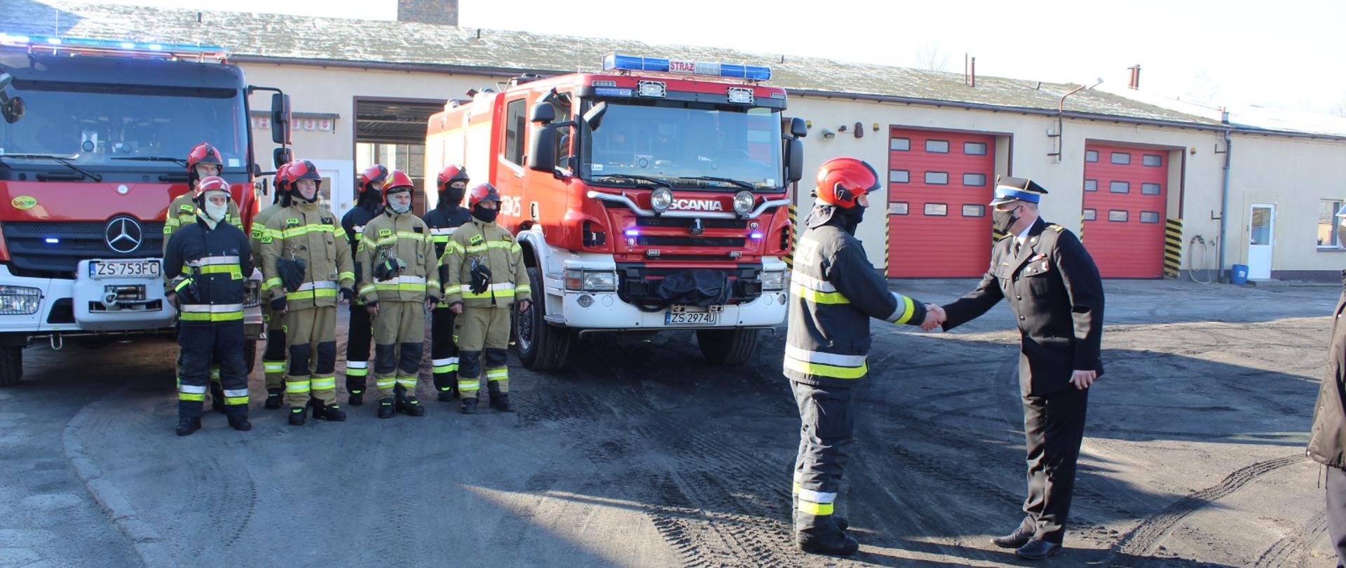 Zdjęcie przedstawia siedmiu strażaków ubranych z strój bojowy stojących przed dwoma wozami strażackimi oraz dwóch strażaków w mundurach wyjściowych. 