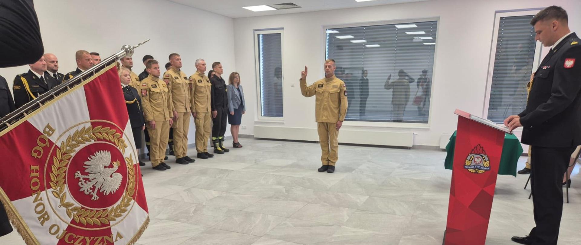 Nowo przyjęty funkcjonariusz stoi na przeciwko sztandaru. Po prawej stronie przy mównicy widoczna sylwetka komendanta miejskiego, na przeciwko niego, w mundurach, w szeregu funkcjonariusze komendy miejskiej i jednostki ratowniczo gaśniczej.