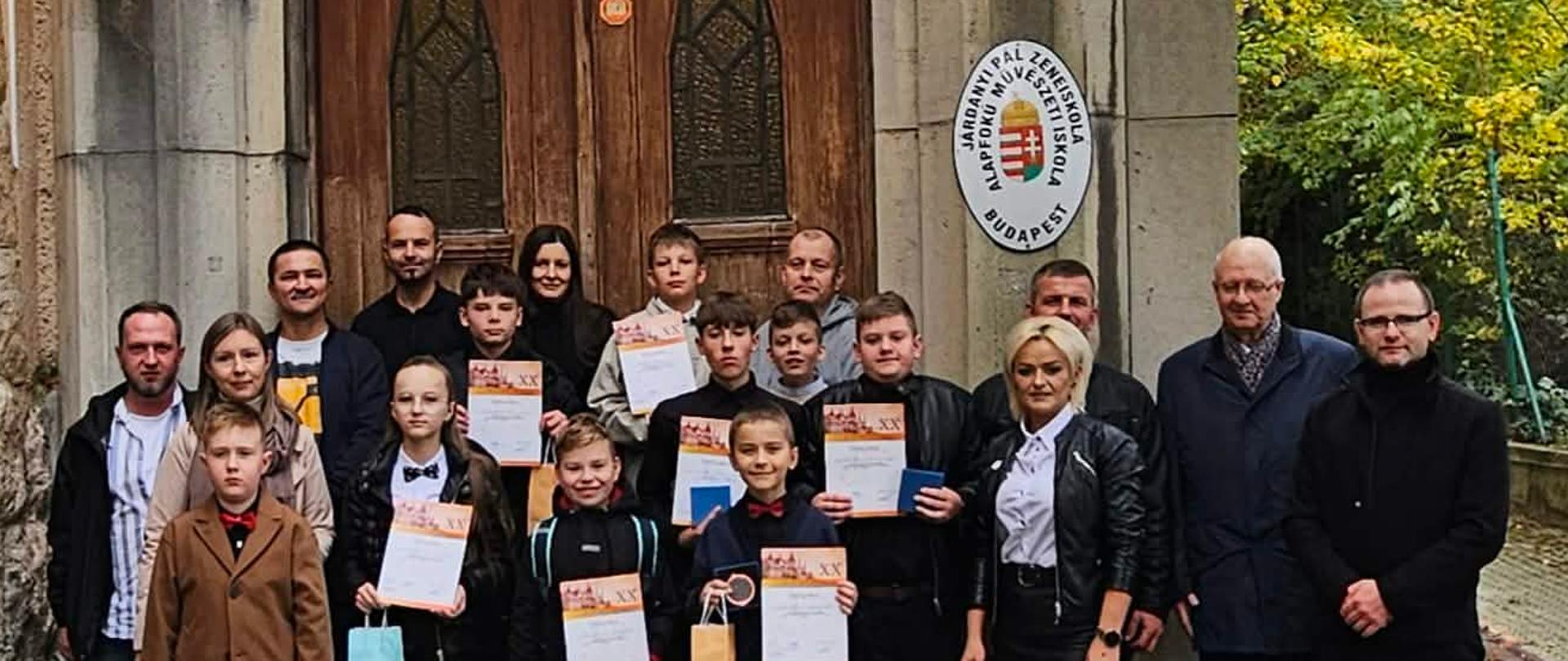 Zdjęcie grupowe wykonane na zewnątrz, przed wejściem do budynku szkoły muzycznej „Járdányi Pál Zeneiskola” w Budapeszcie. Nad ozdobnymi drewnianymi drzwiami widać napis upamiętniający 150-lecie placówki oraz małe flagi Polski i Węgier. Grupa składa się z dzieci – laureatów konkursu trzymających dyplomy i upominki – oraz towarzyszących im dorosłych opiekunów i nauczycieli.