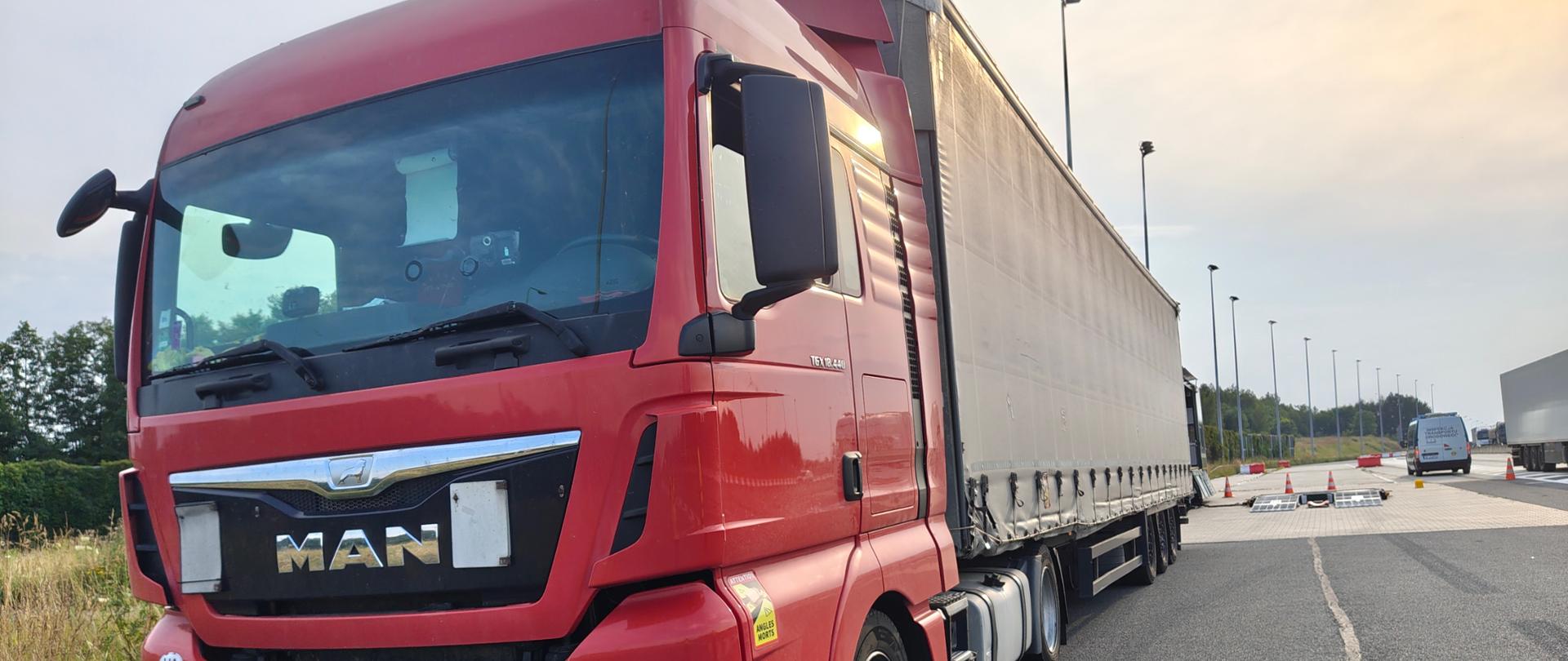 Czerwony pojazd ciężarowy z naczepą, zaparkowany na utwardzonej nawierzchni. Za pojazdem zaparkowany pojazd typu bus z oznaczeniami inspekcji transportu drogowego oraz stanowisko do ważenia pojazdów z pachołkami i najazdami do wag. W tle widoczna infrastruktura drogowa oraz drzewa i krzewy a także inny pojazd ciężarowy.