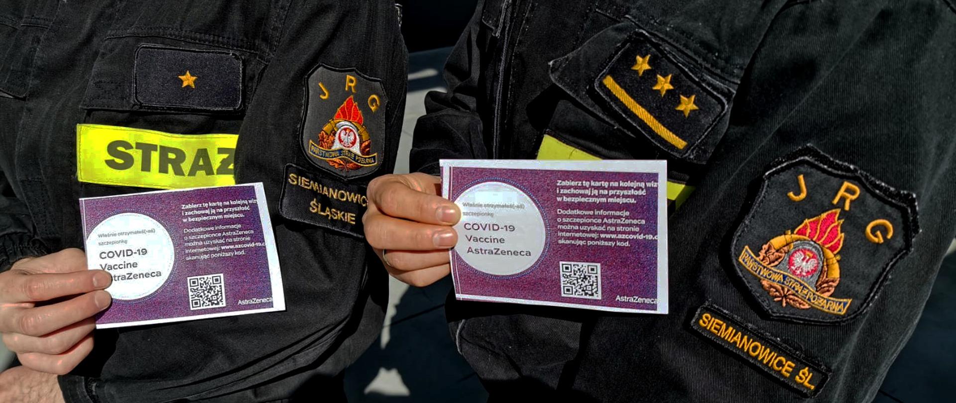 Na zdjęciu widoczne są sylwetki strażaków w ubraniach koszarowych, na ramionach mundurów widoczne są emblematy jednostki ratowniczo-gaśniczej. Strażacy w dłoniach trzymają zaświadczenia o przyjęciu pierwszej dawki szczepionki przeciwko Covid 19