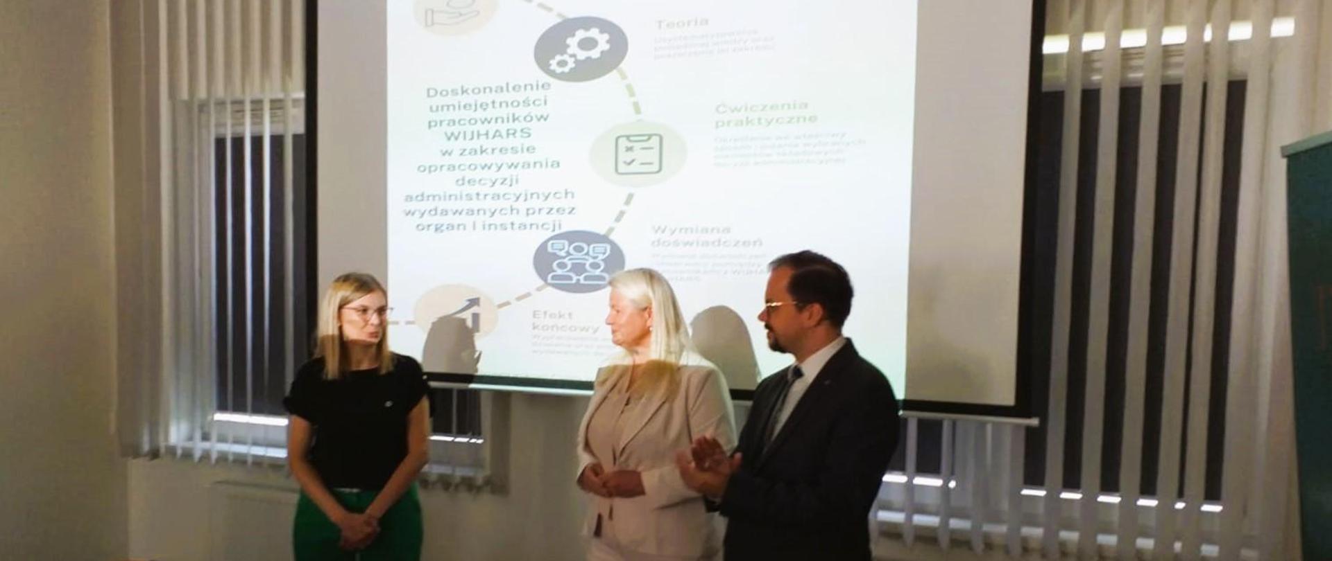 Trzy osoby w sali wykładowej, na tle ekranu z wyświetloną prezentacją