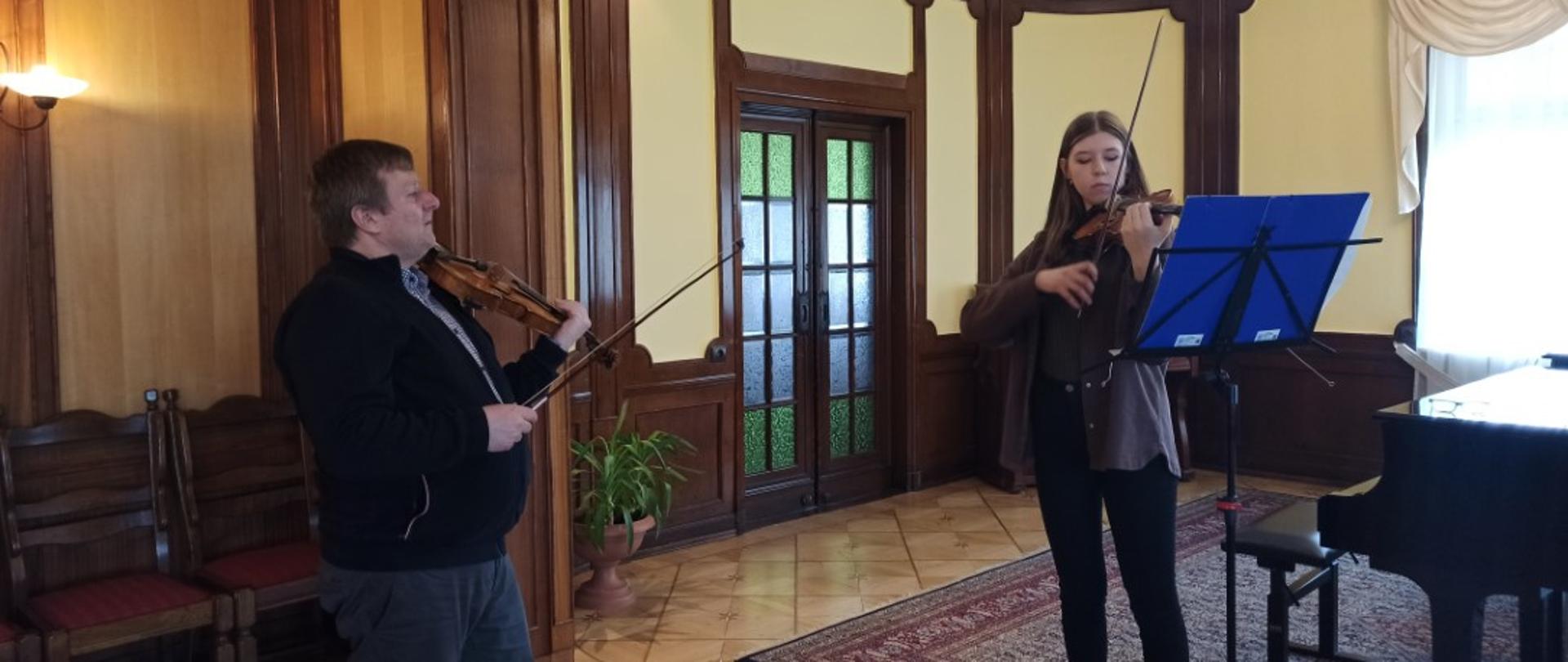 Kolorowe zdjęcie. Uczennica gra na skrzypcach. Po lewej jej stronie stoi prowadzący warsztaty dr hab. Paweł Kukliński. Przed nią znajduje się pulpit z nutami w niebieskiej teczce, a obok fortepian.