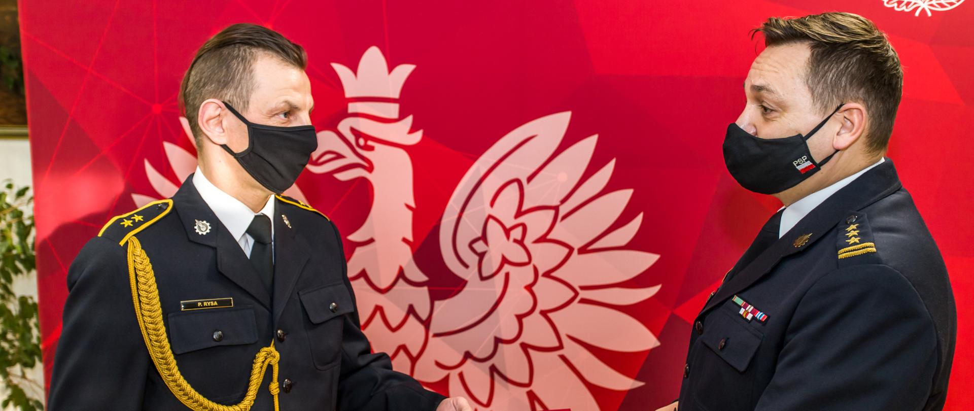 Zdjęcie przedstawia st. bryg. Krzysztofa Kędryka Opolskiego Komendanta Wojewódzkiego PSP (prawa strona kadru) wręczającego lewą ręką ozdobną teczkę z nagrodą Ministra Spraw Wewnętrznych i Administracji st. asp. Rysie, który przyjmuje teczkę do lewej ręki (stoi po lewej stronie kadru). W tle czerwona plansza z białym godłem Polski i białym napisem Państwowa Straż Pożarna Komenda Wojewódzka PSP w Opolu. Obaj strażacy w mundurach galowych PSP z czarnymi maseczkami na twarzach.