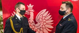 Zdjęcie przedstawia st. bryg. Krzysztofa Kędryka Opolskiego Komendanta Wojewódzkiego PSP (prawa strona kadru) wręczającego lewą ręką ozdobną teczkę z nagrodą Ministra Spraw Wewnętrznych i Administracji st. asp. Rysie, który przyjmuje teczkę do lewej ręki (stoi po lewej stronie kadru). W tle czerwona plansza z białym godłem Polski. Obaj strażacy w mundurach galowych PSP z czarnymi maseczkami na twarzach.