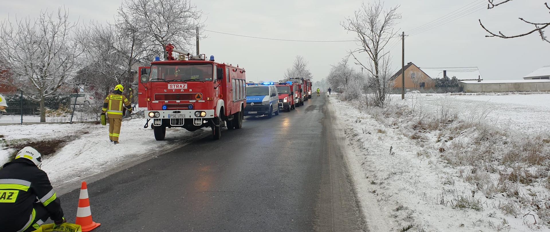 Na zdjęciu strażak przy linii gaśniczej obok pachołka, drugi strażak z wężem gaśniczym, na ulicy wzdłuż ustawione samochody ratowniczo - gaśnicze i radiowóz w oddali strażak kierujący ruchem