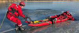 Próbę podjęcia z wody osoby znajdującej się w przeręblu przez dwóch strażaków. Ubrani są oni w suche skafandry. Jeden z nich na końcu przytrzymuje sanie lodowe, drugi znajdujący się na saniach próbuje chwycić osobę poszkodowaną.