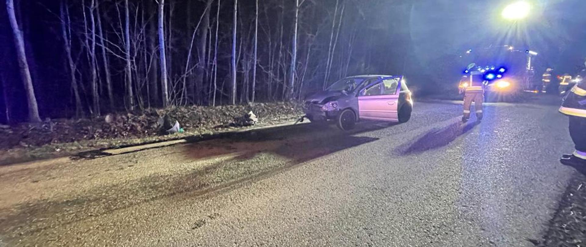 Zdjęcie zrobione nocą. Droga oświetlona przez świtała z samochodu strażackiego. Na drodze stoi uszkodzone srebrne auto.