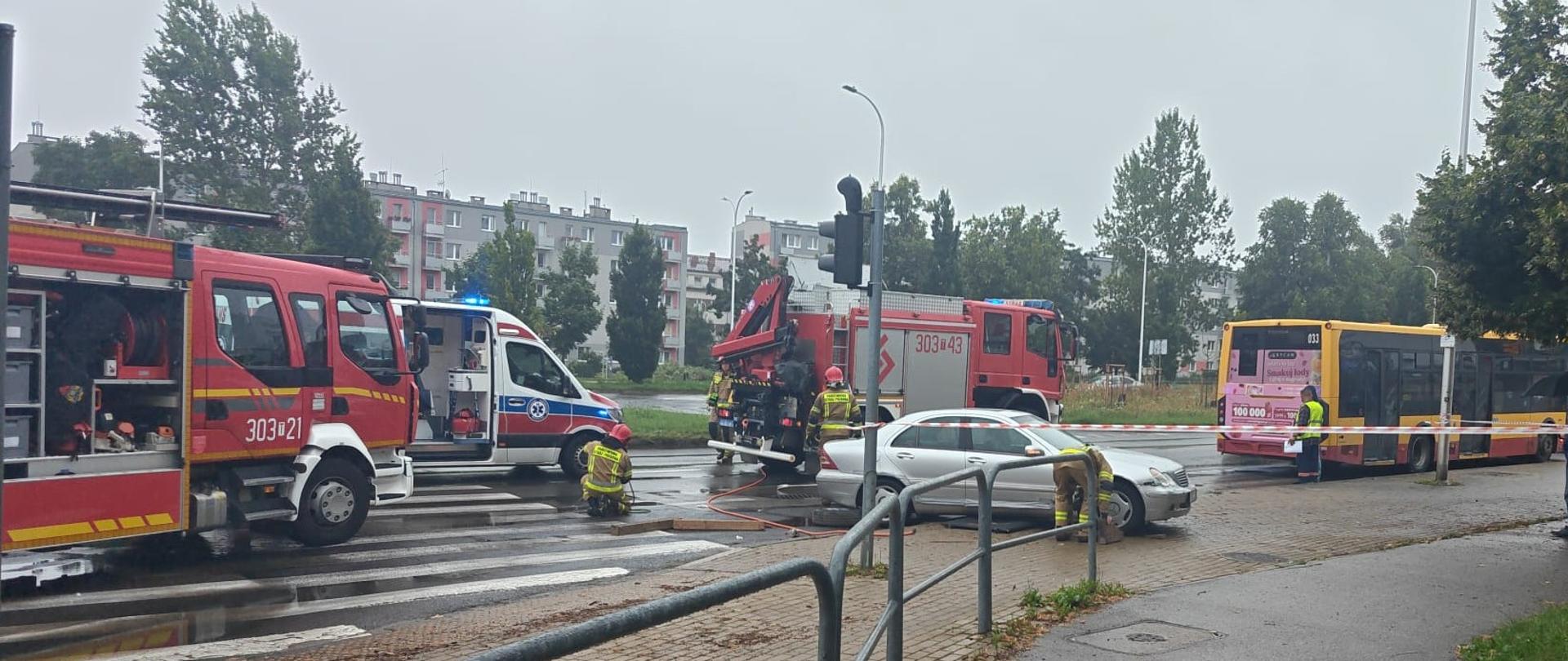 Zdjęcie przedstawia miejsce wypadku: od lewej strony stoją: samochód pożarniczy, ambulans, kolejny samochód strażacki, na jego tle auto osobowe, pod którym znajdował się poszkodowany, a dalej uszkodzony autobus.