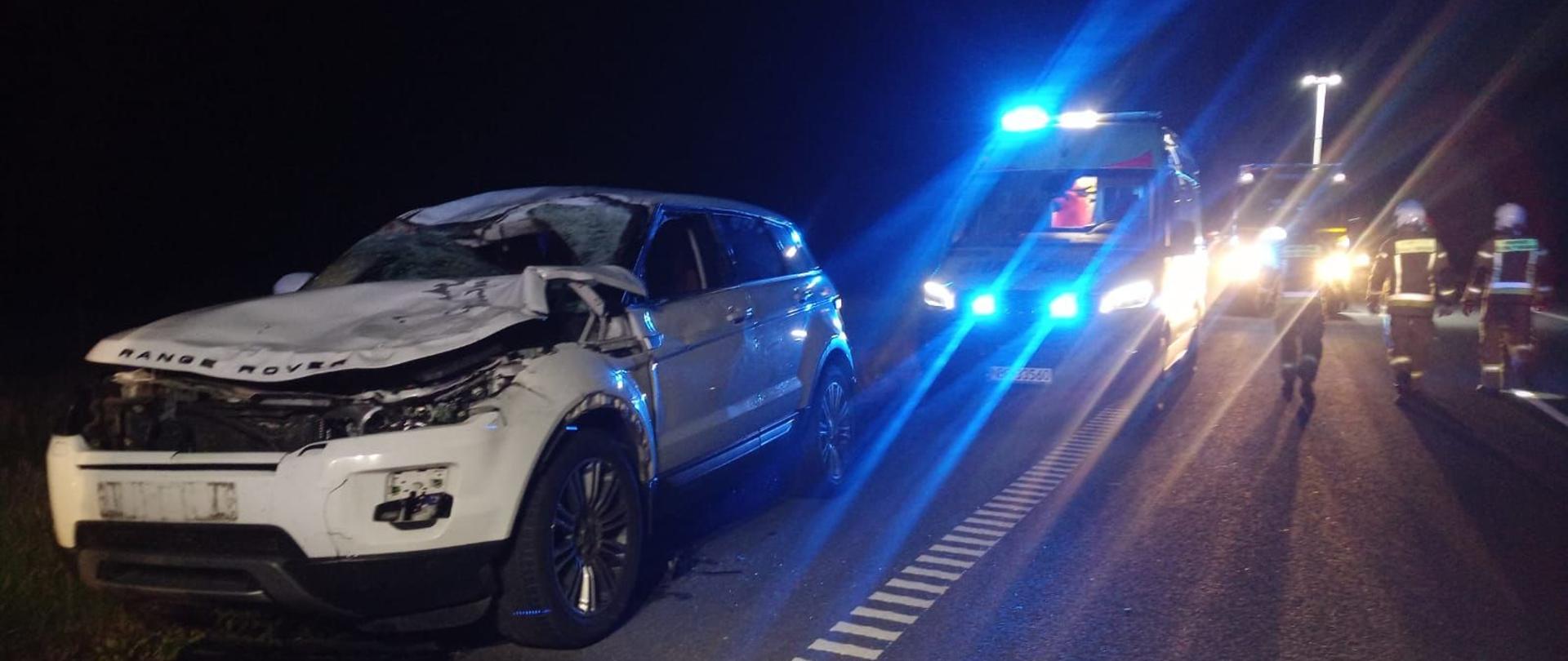 Noc. Z lewej strony rozbite białe auto osobowe. Dalej an drodze pojazdy ratownicze na niebieskich światłach ostrzegawczych. strażacy na drodze.