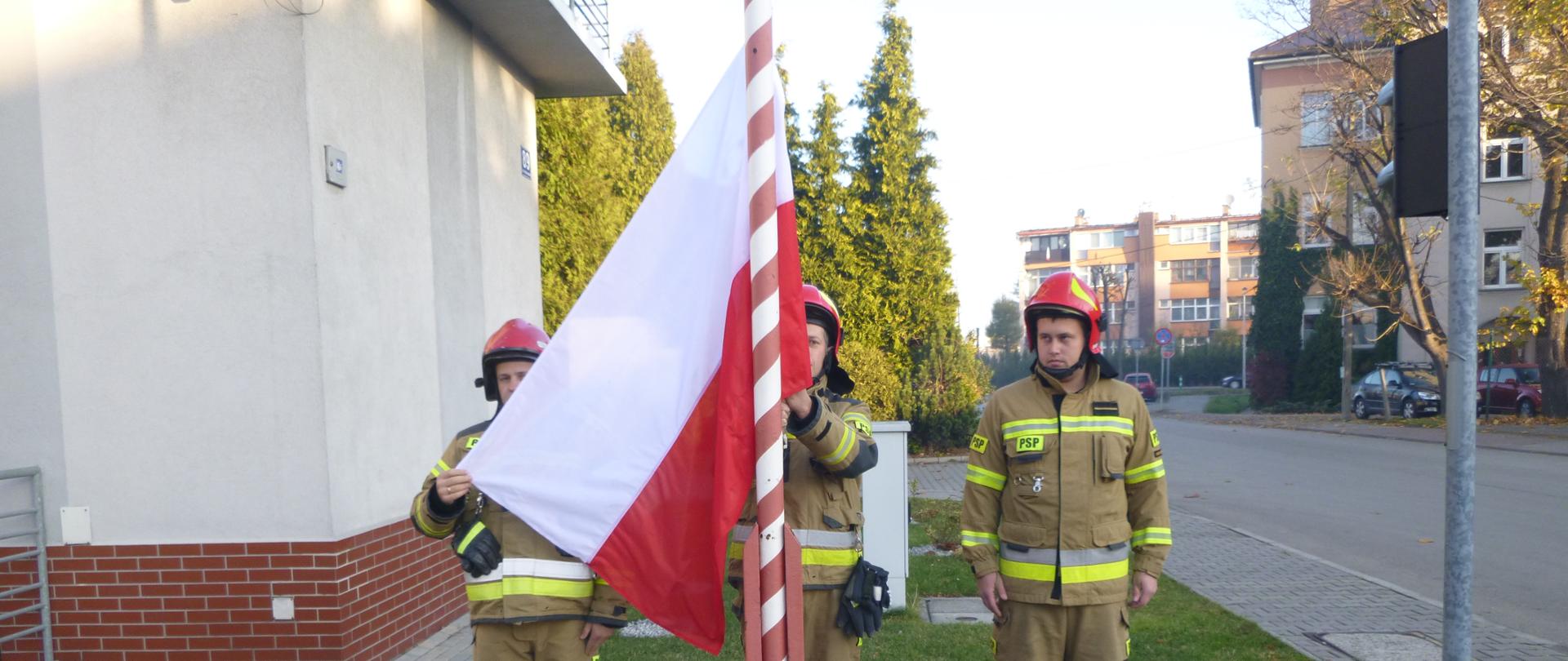 Poczet flagowy w umundurowaniu specjalnym stoi za masztem. Dowódca pocztu podtrzymuje flagę przyczepioną do linki na maszcie.