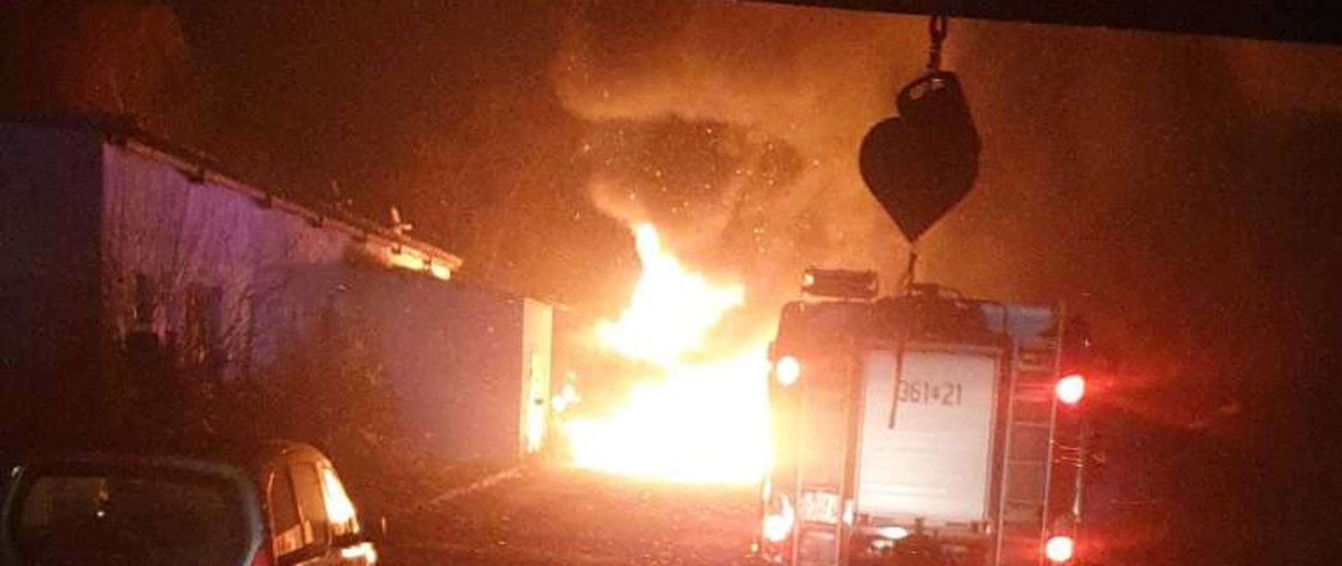 Zdjęcie przedstawia pożar ładowarki kołowej. Zdjęcie zrobione z kabiny samochodu strażackiego podczas dojazdu do miejsca pożaru. Na zdjęciu widać również samochód strażacki i samochód osobowy. Pora nocna.