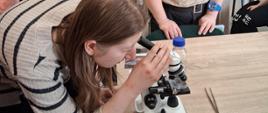 Grupa osób uczestniczących w zajęciach edukacyjnych, zgromadzonych wokół stołu, na którym stoi mikroskop. W centrum uwagi znajduje się osoba pochylona nad mikroskopem, wykonująca obserwacje preparatu. Jej dłonie trzymają korpus urządzenia, a twarz jest skierowana w stronę okularu.
Obok stołu stoi kilka innych osób, uważnie obserwujących przebieg ćwiczenia. Każda z nich ma na ubraniu widoczną plakietkę z imieniem, co sugeruje charakter warsztatowy lub szkoleniowy spotkania. Na blacie, obok mikroskopu, leżą metalowe pęsety oraz niewielka butelka z zakrętką.
W tle widać jasną salę ozdobioną ramkami z kolorowymi ilustracjami przedstawiającymi różne postacie. Dalej stoi rząd ozdobnych figurek przypominających stylizowane postacie zwierząt, ustawionych jedna obok drugiej na ławce. Całość wskazuje na praktyczną część zajęć, podczas której uczestnicy mają możliwość samodzielnego obejrzenia preparatów pod mikroskopem oraz poszerzenia wiedzy o tematy związane z ochroną zdrowia.
