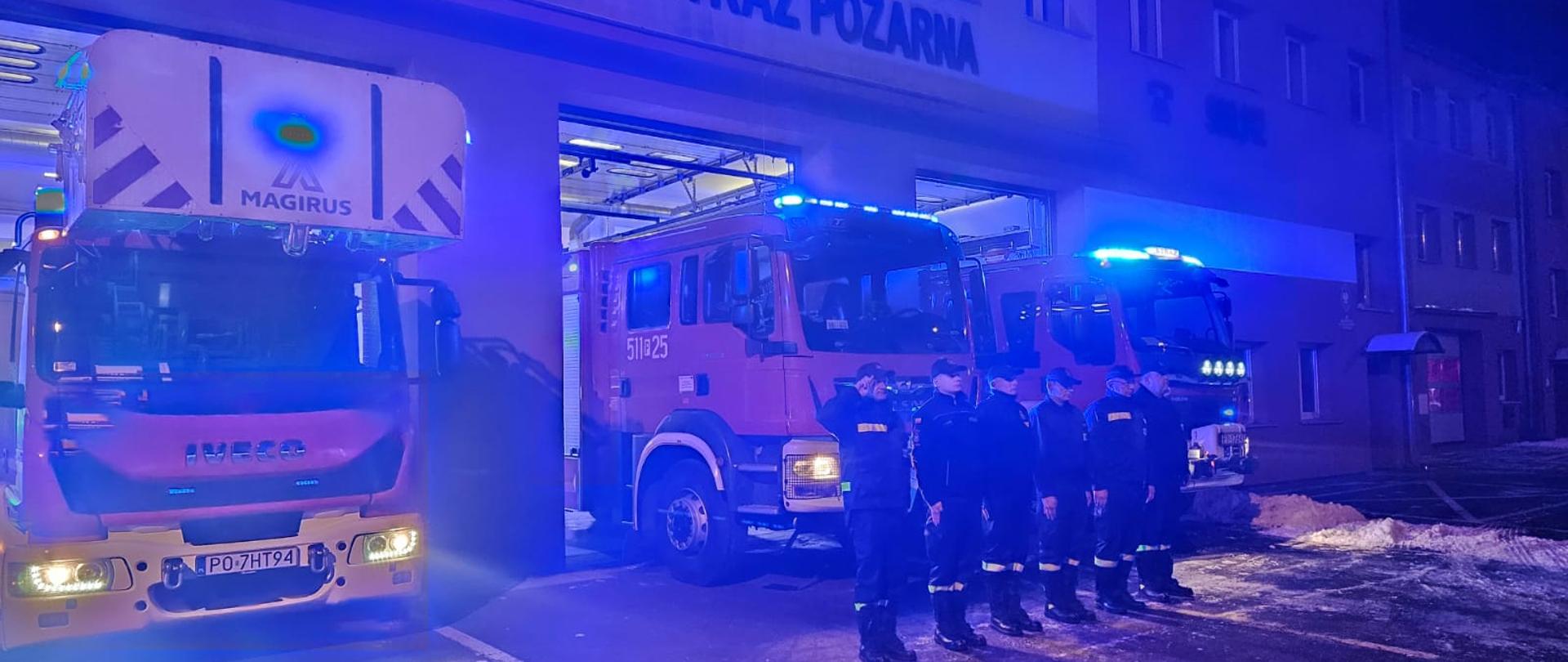 Sześciu strażaków stojących w szeregu na tle samochodów bojowych z włączonymi światłami alarmowymi.