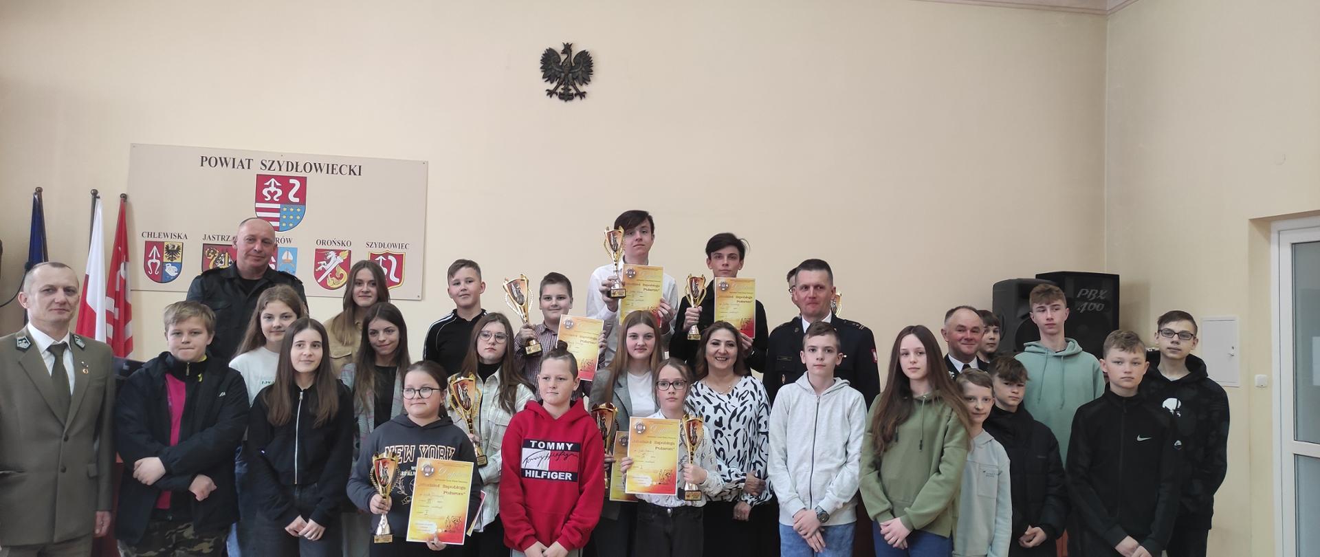 Fotografia przedstawiająca finalistów Powiatowego Turnieju Wiedzy Pożarniczej wraz z Wicestarostą powiatu szydłowieckiego, przedstawicielem Regionalnej Dyrekcji Lasów Państwowych w Radomiu, Komendantem Powiatowym PSP w Szydłowcu oraz Zastępcę Komendanta Powiatowego PSP w Szydłowcu.