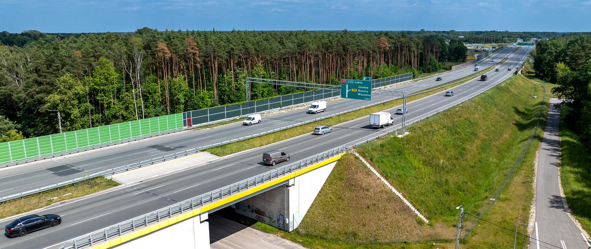Dwujezdniowa droga ekspresowa z umiarkowanym ruchem samochodów, słoneczna pogoda z delikatnym zachmurzeniem. Po obu stronach drogi las.
