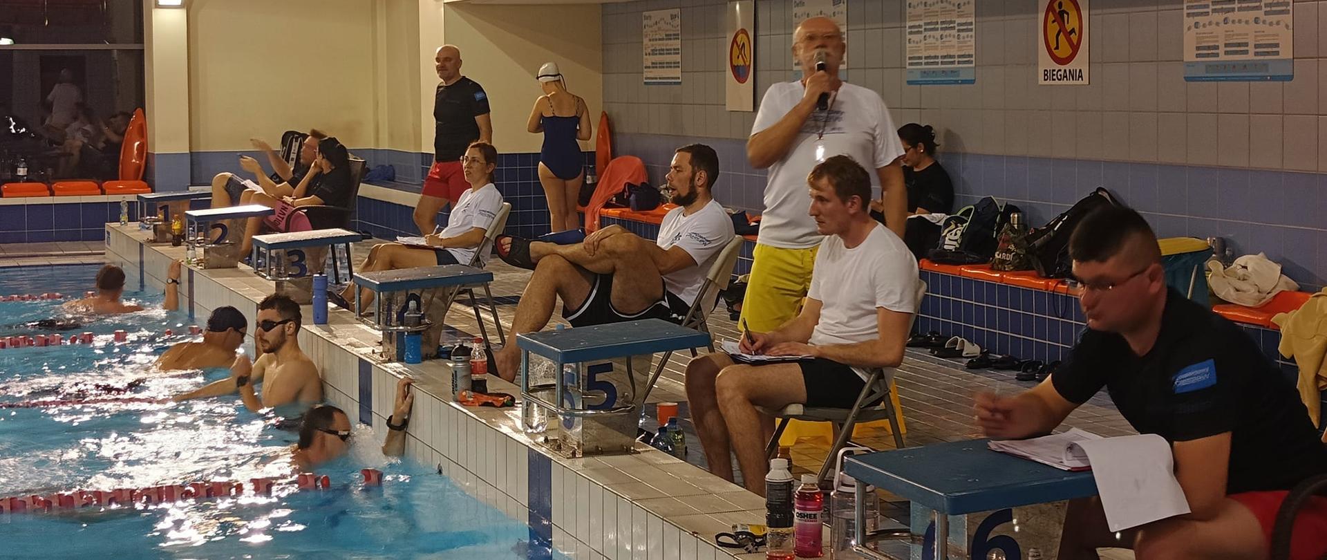 Basen, na miejscach startowych siedzą mężczyźni w strojach kąpielowych zapisujący karty z wynikami, jeden z nich trzyma mikrofom, w basenie widoczni pływacy