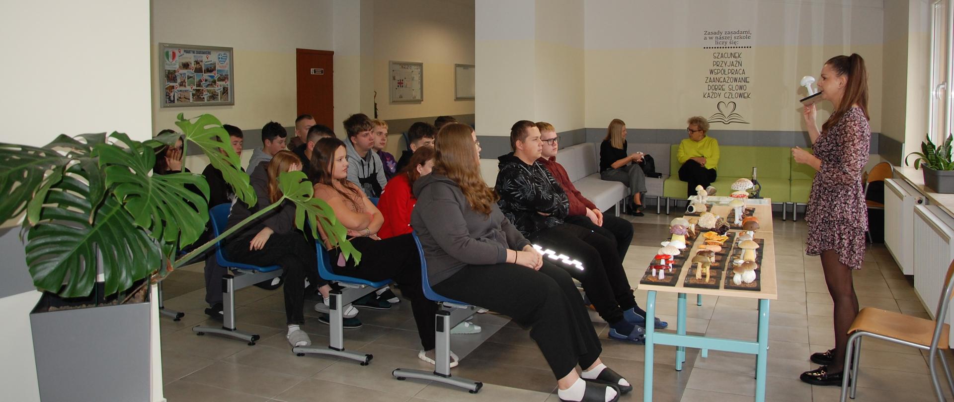 Młodzież siedzi przed wystawą grzybów słuchając wykładu kobiety, kobieta stoi przed nimi trzymając model grzyba