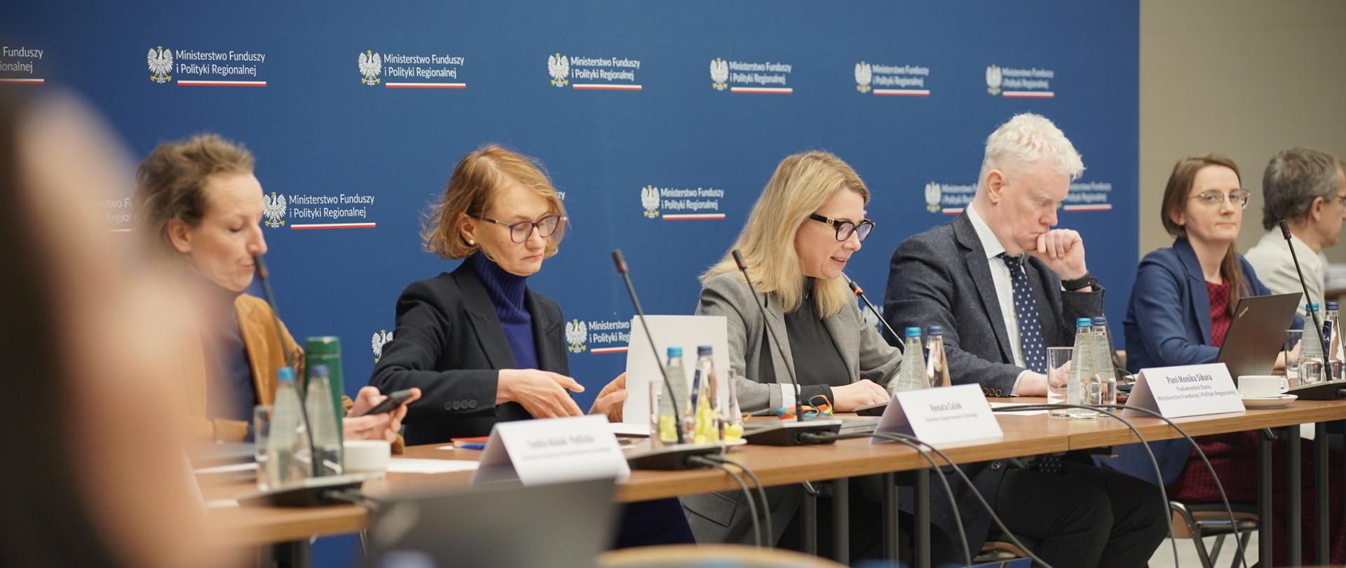 Pięć osób siedzi obok siebie przy stole. Wśród nich w środku siedzi wiceminister Monika Sikora i mówi do mikrofonu.