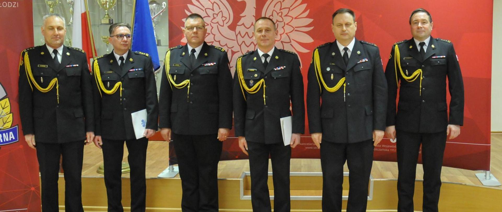 Na zdjęciu widać szczęściu strażaków w ciemnych mundurach strażackich. Dwóch z nich trzyma w lewym ręku białe teczki. Wszyscy pozują do zdjęcia. W tle są fagi Polski i Unii Europejskiej oraz banery. Zdjęcie zrobione jest w pomieszczeniu.