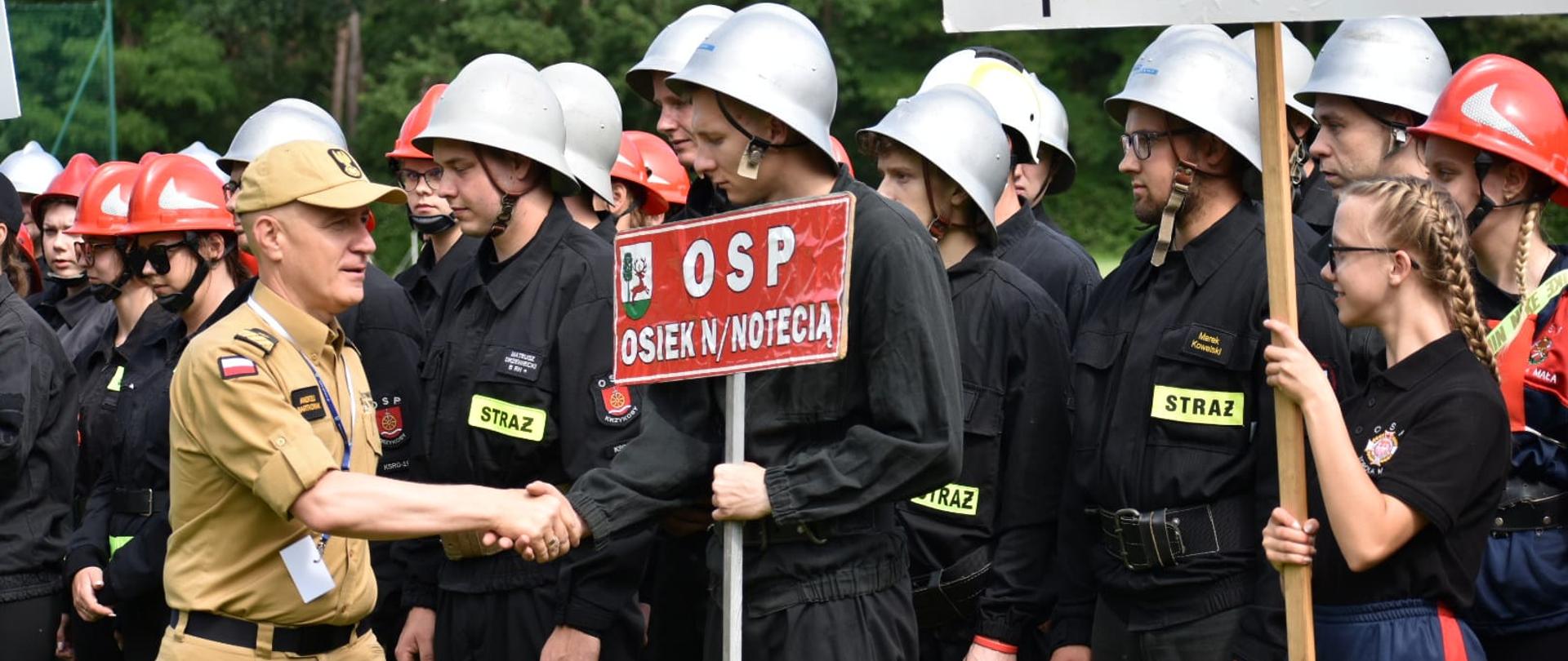 Komendant główny PSP wita się z druhami OSP, jeden z nich trzyma tablicę z nazwą jednostki OSP Osiek n/Notecią
