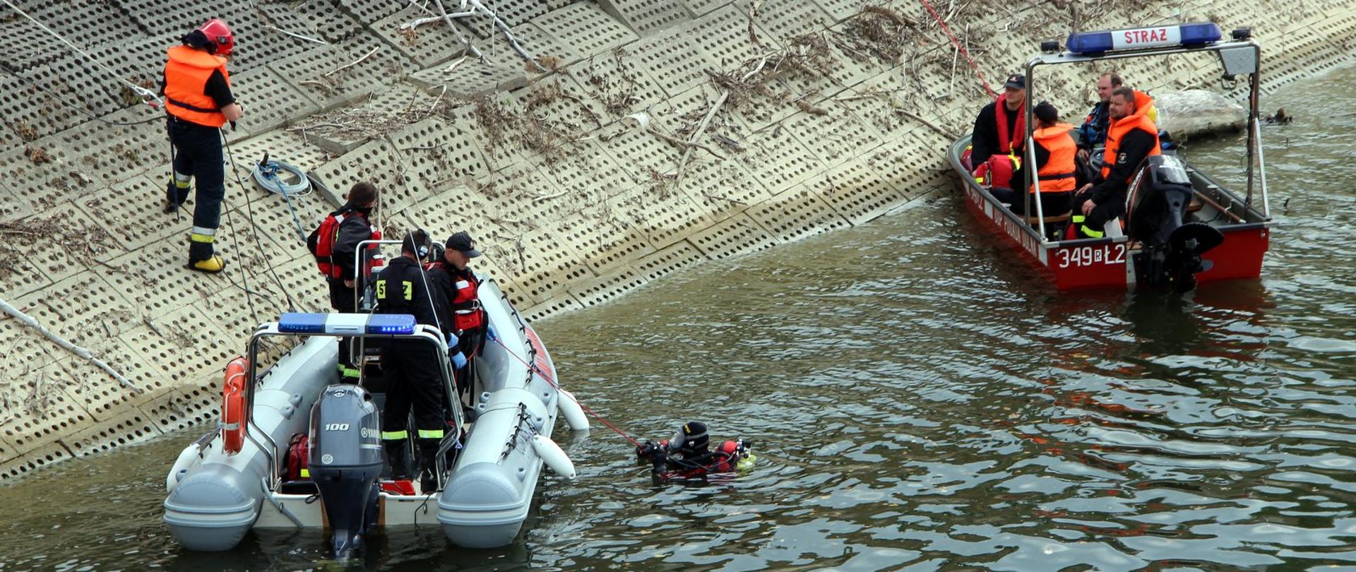Zdjęcie wykonane na zewnątrz, przy zbiorniku wodnym. Na fotografii widać dwie łodzie strażackie, pomiędzy nimi w wodzie zanurzony jest strażak płetwonurek. 