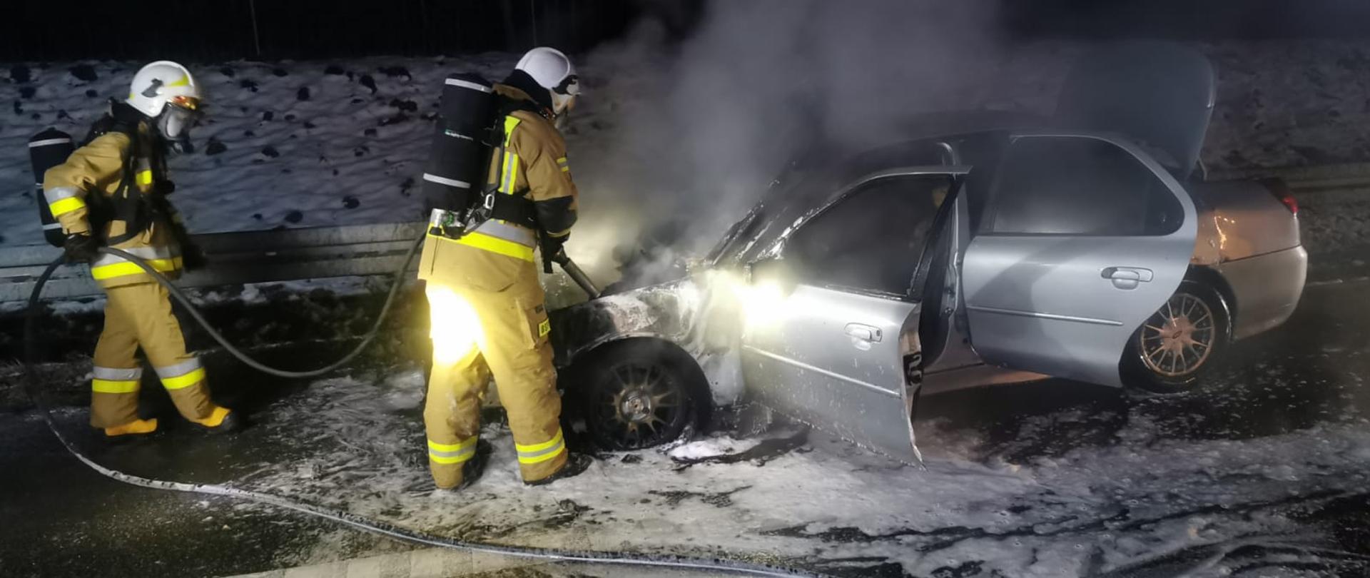 Druhowie OSP ubrani w ubrania specjalne i aparaty ochrony układu oddechowego gaszą palący się samochód osobowy.
