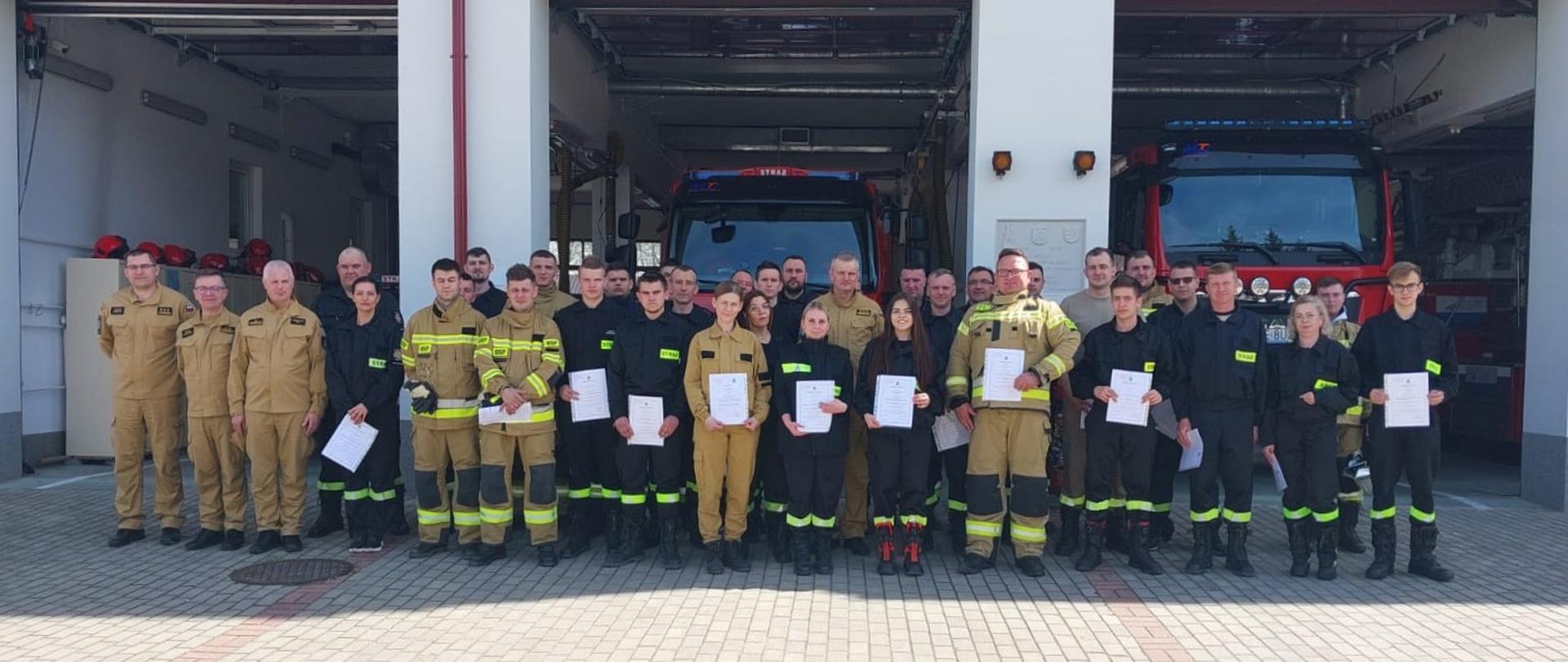 Zdjęcie przedstawia grupę ponad 30 strażaków, stojących na zewnątrz remizy strażackiej w słoneczny dzień. Strażacy są ustawieni w kilku rzędach przed otwartymi bramami garażowymi, za którymi widać zaparkowane wozy strażackie. Większość z nich ubrana jest w ciemne mundury bojowe, niektórzy mają na sobie jasne, robocze kombinezony. Wszyscy trzymają w rękach białe dokumenty, prawdopodobnie zaświadczenia, co sugeruje uroczystość zakończenia szkolenia. Nad ich głowami, na ścianie budynku, widnieje czerwony napis "PAŃSTWOWA STRAŻ POŻARNA".