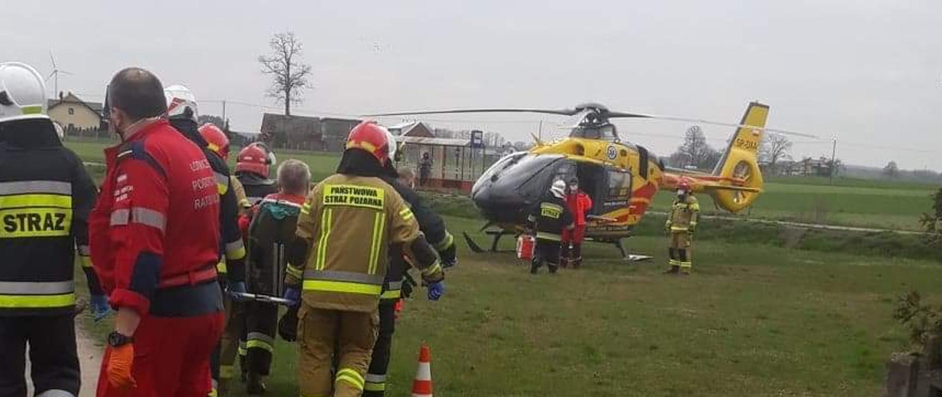 Na zdjęciu strażacy i ratownicy transportują osobę do śmigłowca Lotniczego Pogotowia Ratunkowego. Na pierwszym planie osoby ubrane w umundurowanie specjalne obrócone tyłem do fotografa. Na drugim planie statek powietrzny znajdujący się na trawniku przy posesji.