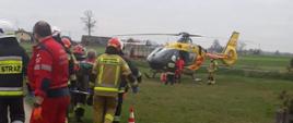 Na zdjęciu strażacy i ratownicy transportują osobę do śmigłowca Lotniczego Pogotowia Ratunkowego. Na pierwszym planie osoby ubrane w umundurowanie specjalne obrócone tyłem do fotografa. Na drugim planie statek powietrzny znajdujący się na trawniku przy posesji.