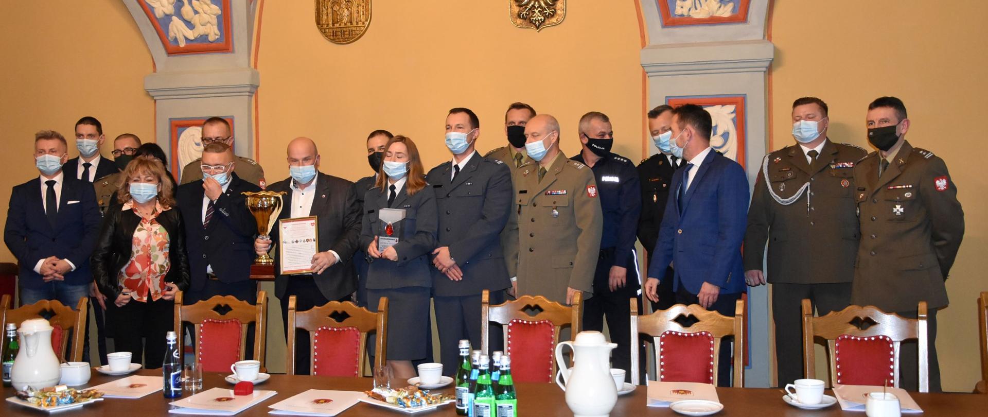 Zdjęcie przedstawia grupę osób na spotkaniu wewnątrz zabytkowego pomieszczenia. Funkcjonariusze w mundurach wyjściowych i osoby cywilne stoją w dwuszeregu i pozują do zdjęcia. W tle zabytkowe wykończenia sklepień łukowych.