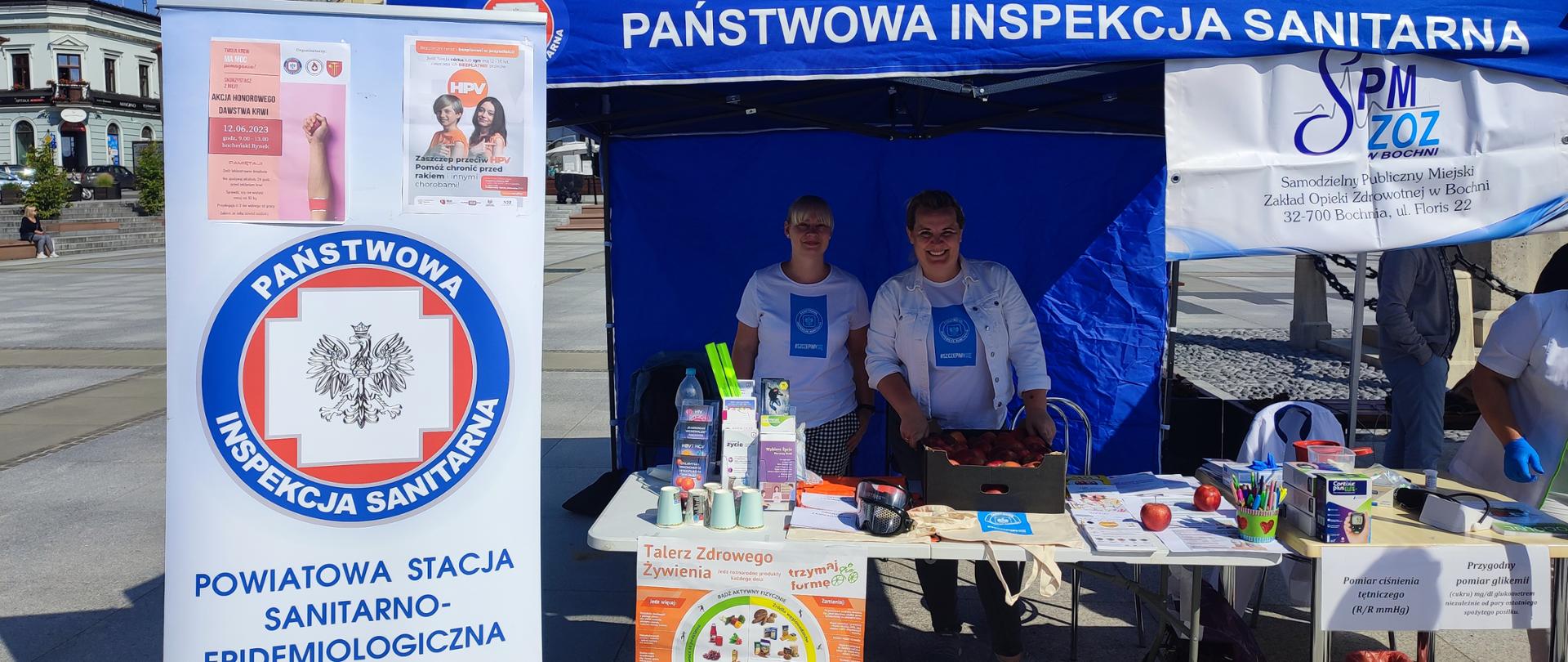 Po lewej rollup z logo Państwowej Inspekcji Sanitarnej, na środku niebieski namiot, pod namiotem dwie kobiety i stolik z ulotkami