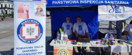 Po lewej rollup z logo Państwowej Inspekcji Sanitarnej, na środku niebieski namiot, pod namiotem dwie kobiety i stolik z ulotkami
