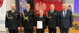 Na zdjęciu widać pięć osób, czterech mężczyzn i jedną kobietę. Osoby stoją w jednym rzędzie i pozują do zdjęcia. Pani ubrana jest w strój galowy członka ochotniczej straży pożarnej i trzyma w ręku białą teczkę. Po jej prawej stronie stoją dwaj mężczyźni w galowych mundurach strażaka Państwowej Straży Pożarnej. Jeden z nich ma okulary. Z drugiej strony stoją też dwaj mężczyźni. Jeden ubrany jest w strój galowy strażaka Państwowej Straży Pożarnej a jeden w ciemny garnitur. W tle widać banery Państwowej Straży Pożarnej i ochotniczej Straży pożarnej. Zdjęcie zrobione jest w pomieszczeniu.