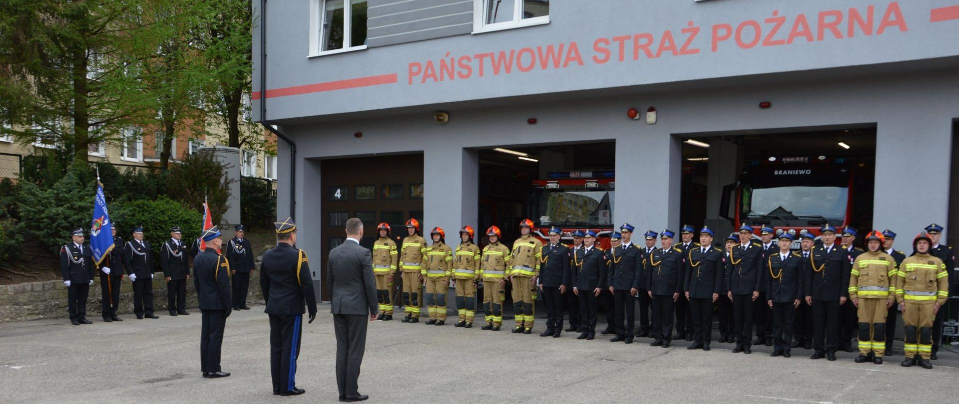 Plac wewnętrzny. Wzdłuż budynku strażnicy strażacy ustawieni w szeregu w mundurach galowych i bojowych, za nimi w bramach garażowych samochody strażackie. Z lewej strony sztandary.