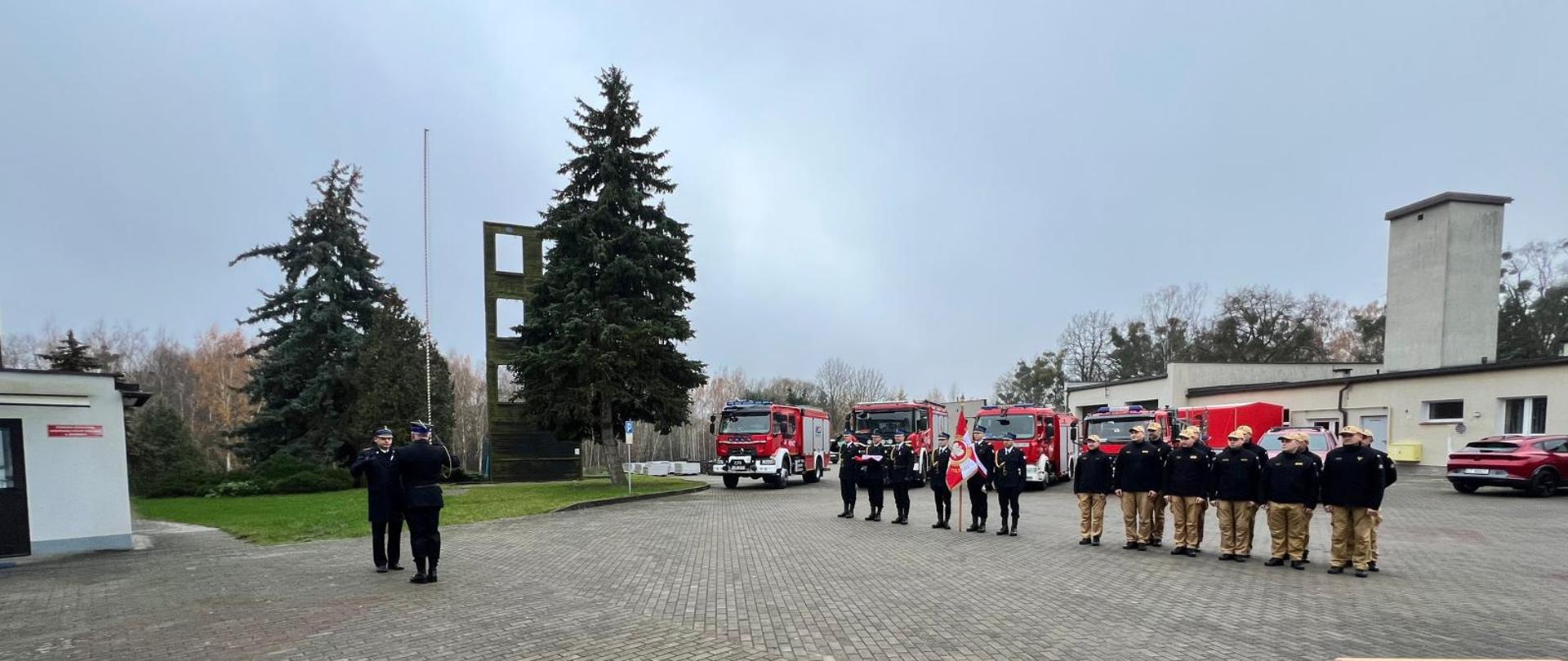 Zdjęcie przedstawia apel podczas Narodowego Święta Niepodległości. Na placu komendy stoją w dwuszeregu strażacy w mundurach służbowych. Obok nich stoi poczet flagowy i sztandarowy Komendy. Po lewo prowadzący uroczystość składa meldunek Komendantowi. W oddali wspinalnia straży i dwie duże choinki.