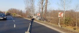 Droga, po której jedzie samochód osobowy, z prawej strony, ciężarówka w rowie, w tle strażacy i karetka pogotowia ratunkowego. W prawej części zdjęcia młody las.