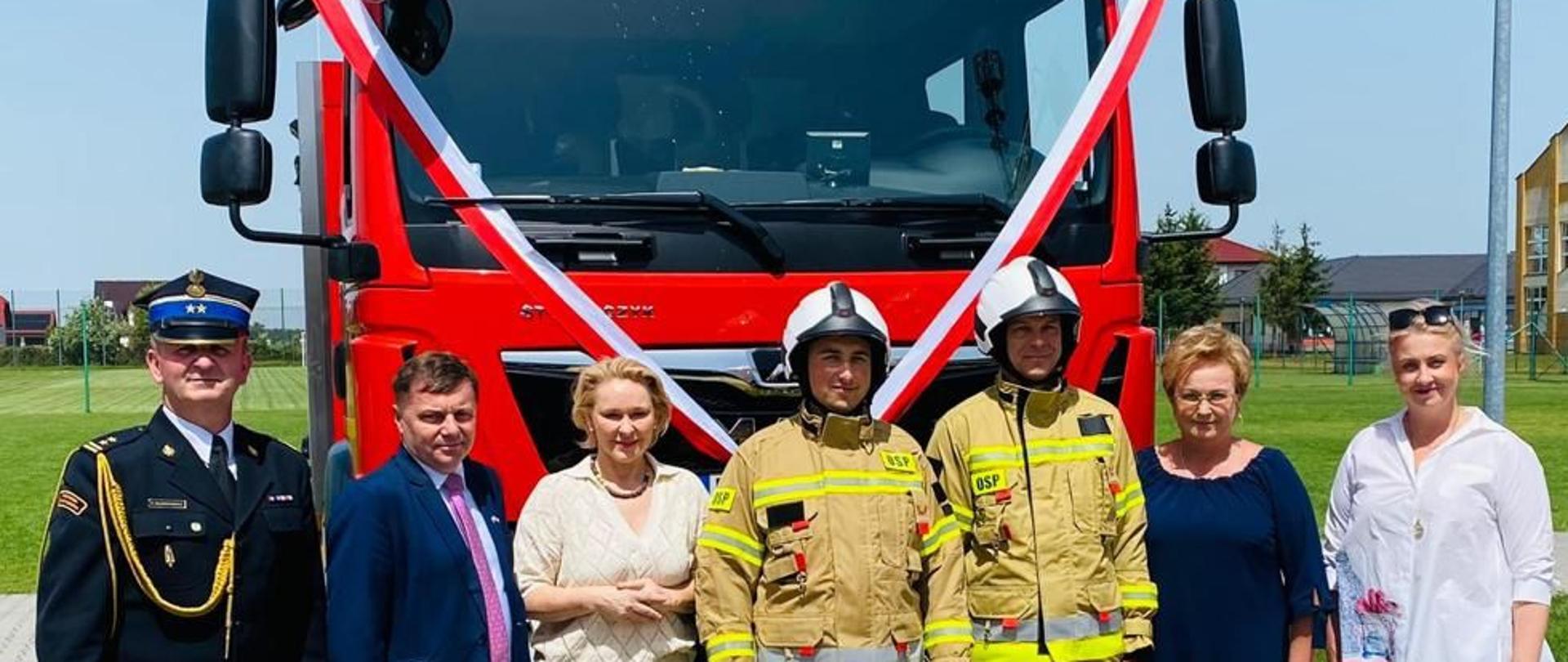 Zaproszeni goście stoją z strażakami przed wozem strażackim. W tle boisko do piłki nożnej.