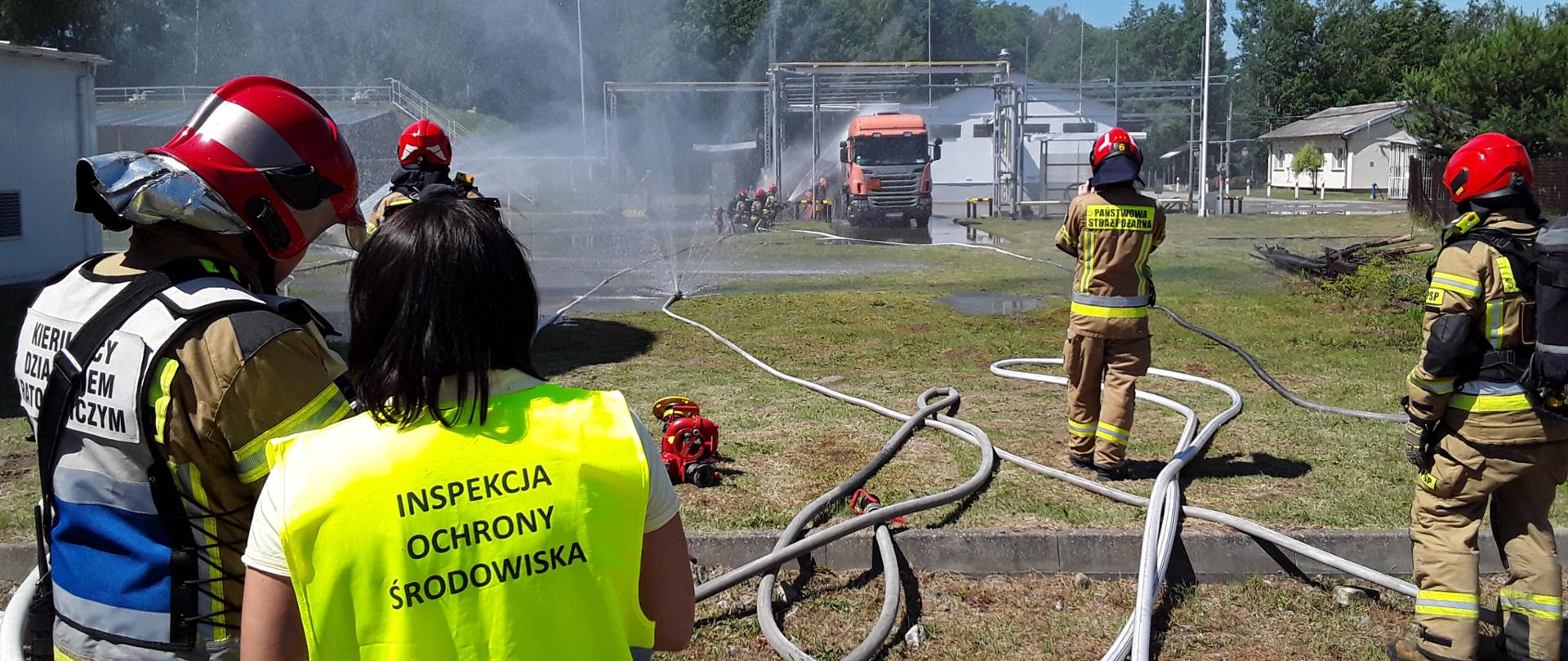 Inspektor WIOŚ w Warszawie wraz z grupą funkcjonariuszy Państwowej Straży Pożarnej nadzoruje spryskiwanie autocysterny wodą przez drugą grupę strażaków. Nadzorujący stoją w znacznej odległości od cysterny. Na trawie porozciągane są liczne węże strażackie, niektóre podłączone do zraszaczy spryskujących cały teren. Teren otaczają budynki zakładowe i drzewa.