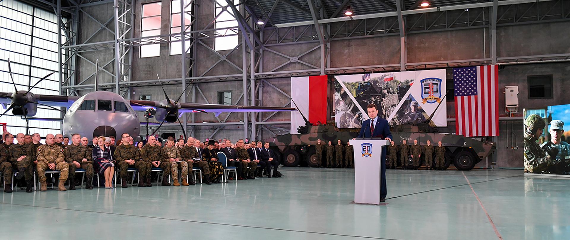 Minister Mariusz Błaszczak przy mównicy. Odprawa ws. ćwiczenia Defender Europe 20, w tle żołnierze i pojazdy opancerzone. Flagi PL i USA.