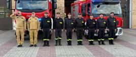 
Dziewięciu strażaków stoi przed remizą. Za nimi zaparkowane są dwa wozy strażackie. Dwóch strażaków z lewej strony ma piaskowe mundury, a pozostałych siedmiu jest ubranych w czarne stroje. Strażak na lewym końcu grupy salutuje.