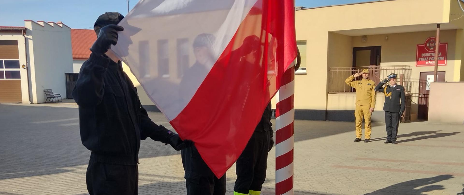 Zdjęcie przedstawia moment podniesienia flagi państwowej na maszt, w tle strażacy i kierownictwo Komendy oddają hołd