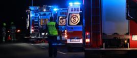 Dwa wozy strażackie oraz 2 karetki pogotowia stoją na jezdni z włączonymi sygnałami błyskowymi podczas zdarzenia. Na drodze stoi również policjant oraz druhowie OSP.