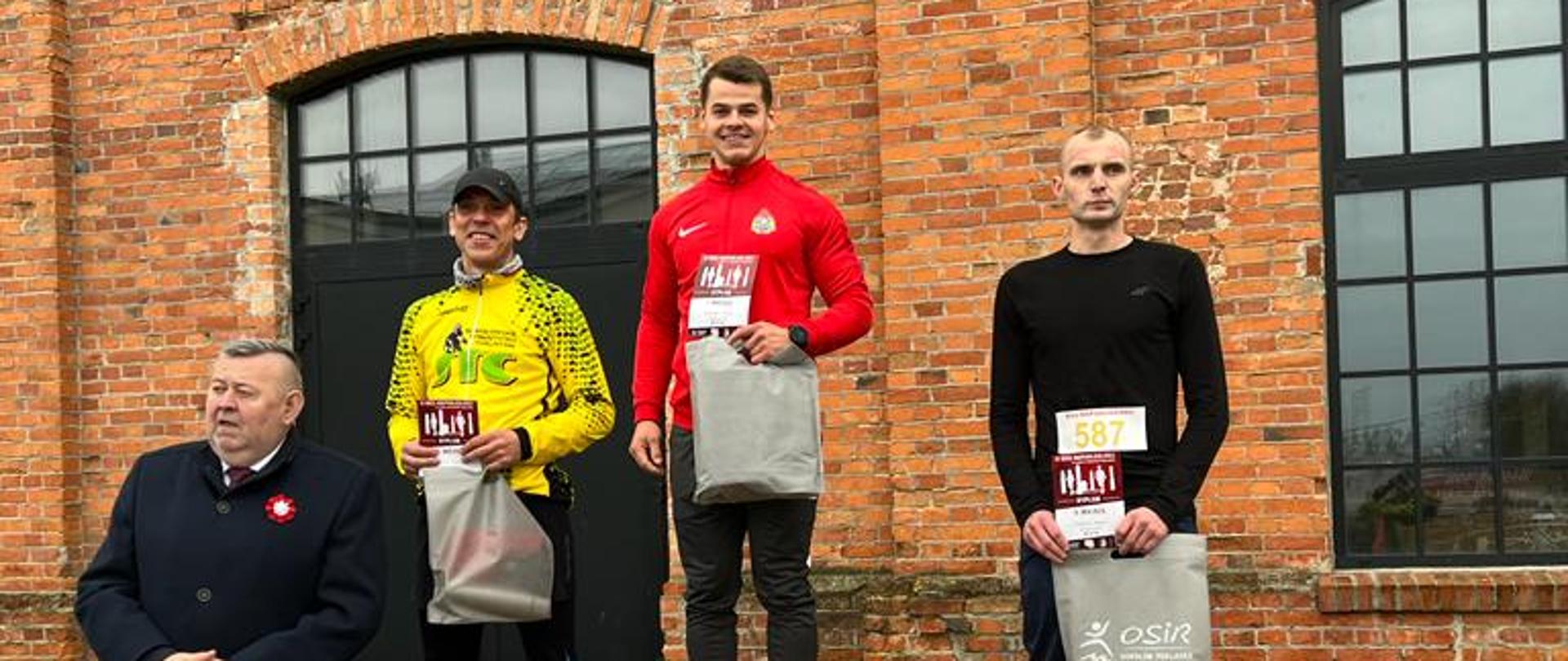 Zwycięstwo asp. Piotra Domańskiego w 11. Bieg Niepodległości - pod budynkiem stoją zawodnicy w dresach w rękach mają dyplomy i nagrody, 