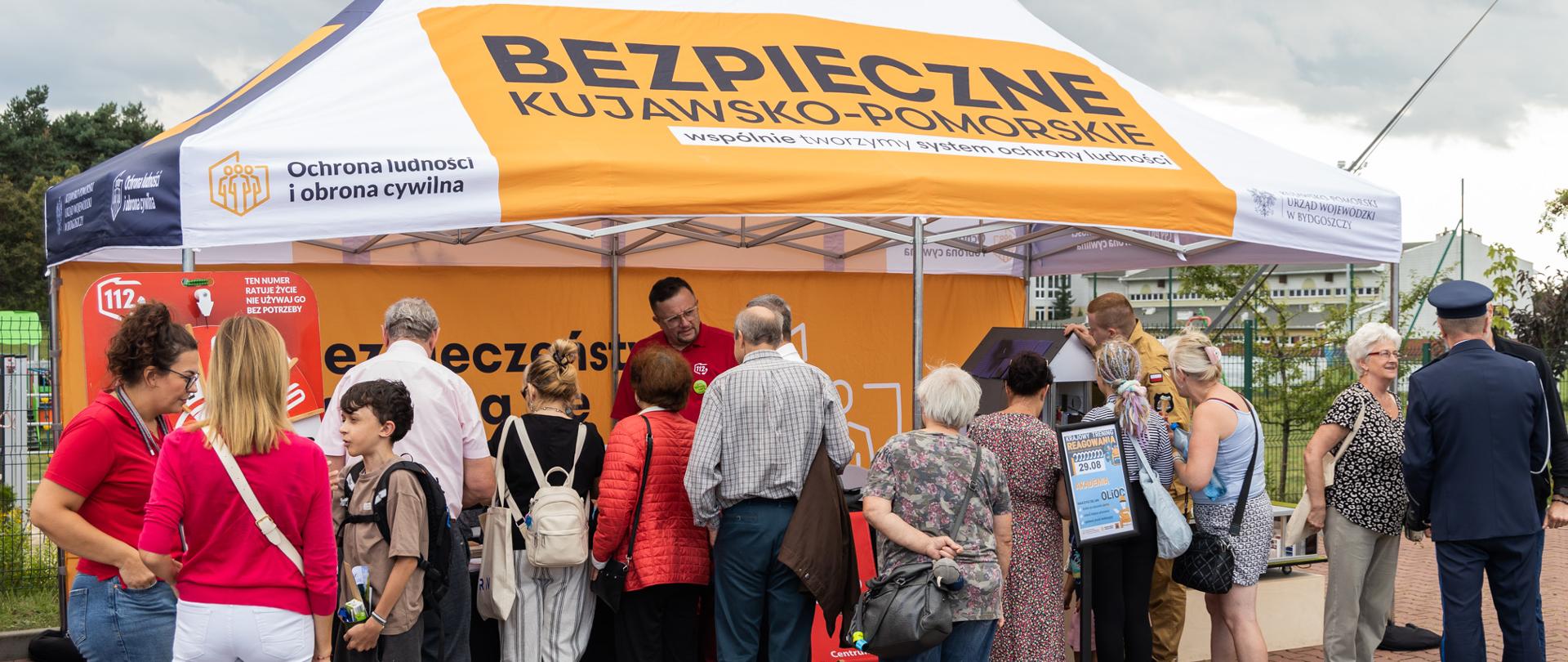 Liczna grupa ludzi zgromadziła się pod dużym, biało-pomarańczowym namiotem z napisem „BEZPIECZNE KUJAWSKO-POMORSKIE”. Zwiedzający rozmawiają z osobami obsługującymi stoisko oraz przeglądają materiały informacyjne. Po prawej stronie stoiska stoi strażak w ciemnym mundurze galowym.
