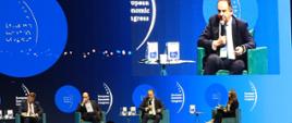 Wiceminister rozwoju i technologii Andrzej Gut-Mostowy podczas panelu "Turystyka - nowe otwarcie" na Europejskim Kongresie Gospodarczym w Katowicach, A. Gut-Mostowy siedzi w fotelu i trzyma w ręce mikrofon, w górnym prawym rogu telebim przedstawiający przemawiającego wiceministra