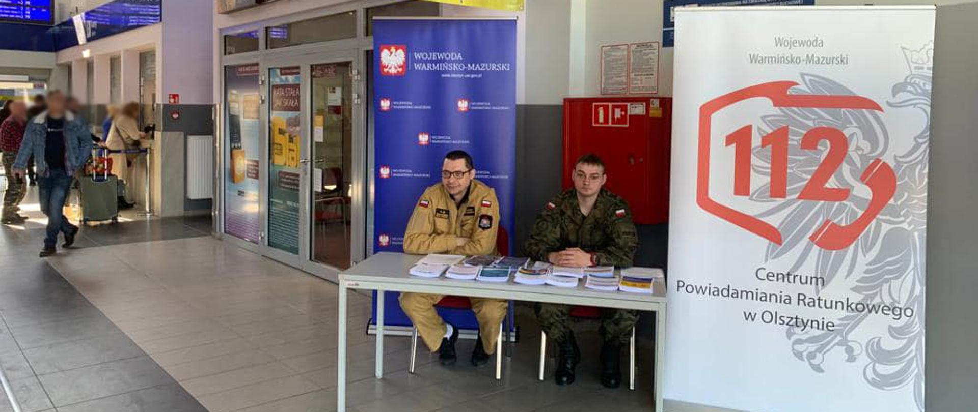 Punkt informacyjny dla uchodźców z Ukrainy, na dworcu kolejowym w Elblągu. Przy stoliku siedzi strażak państwowej straży pożarnej i żołnierz wojsk obrony terytorialnej. Na stoliku zgromadzone są materiały informacyjne, broszury i ulotki.
